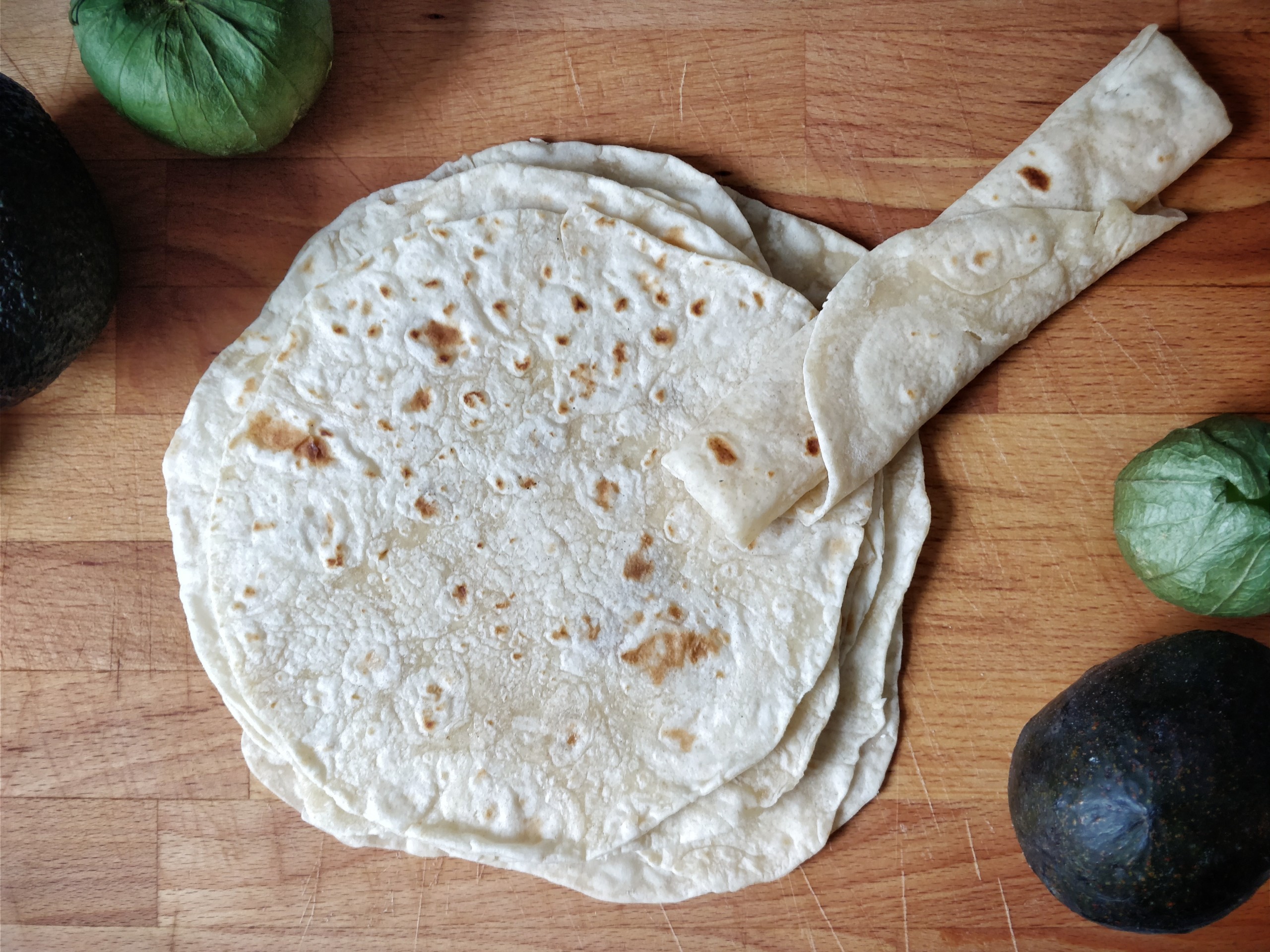 A stack of freshly made tortillas on a wooden board, with some avocados and tomatillos placed around. One tortilla is rolled up to indicate softness.