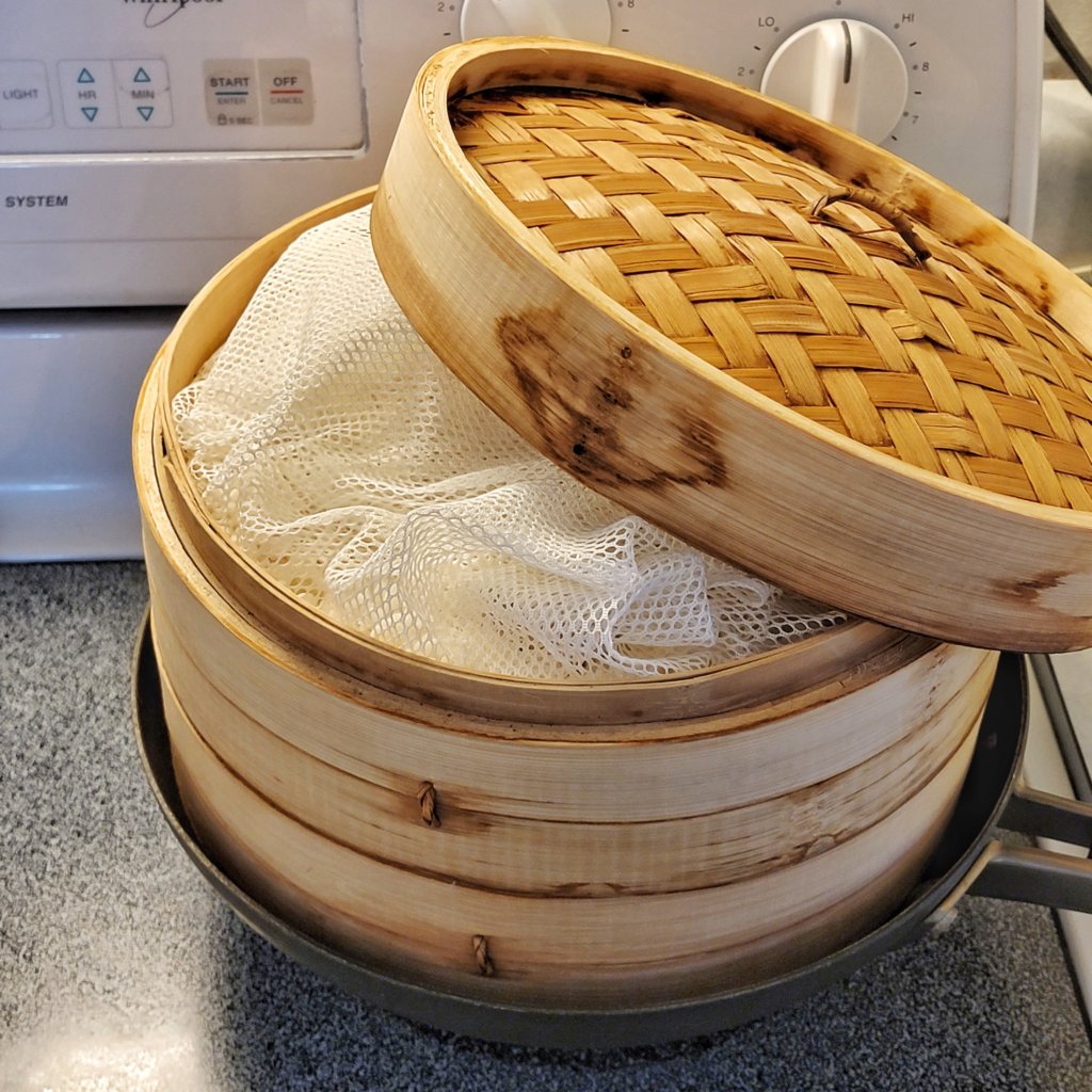 The two bamboo steamer layers are set in a wok on the stove. The lid is open showing that the nylon mesh is folded over top of the rice.