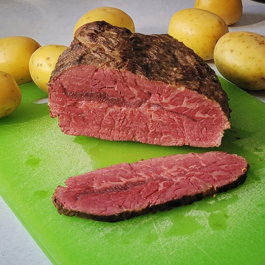 Corned beef on a cutting board sliced to reveal a cross-section of pink and well-marbled beef. Yukon Gold potatoes are scattered behind.