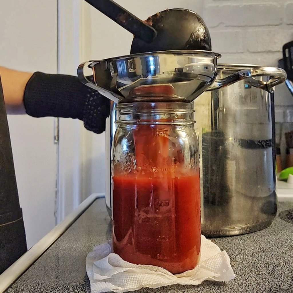Ladling tomato sauce into a jar with a stainless steel canning funnel.
