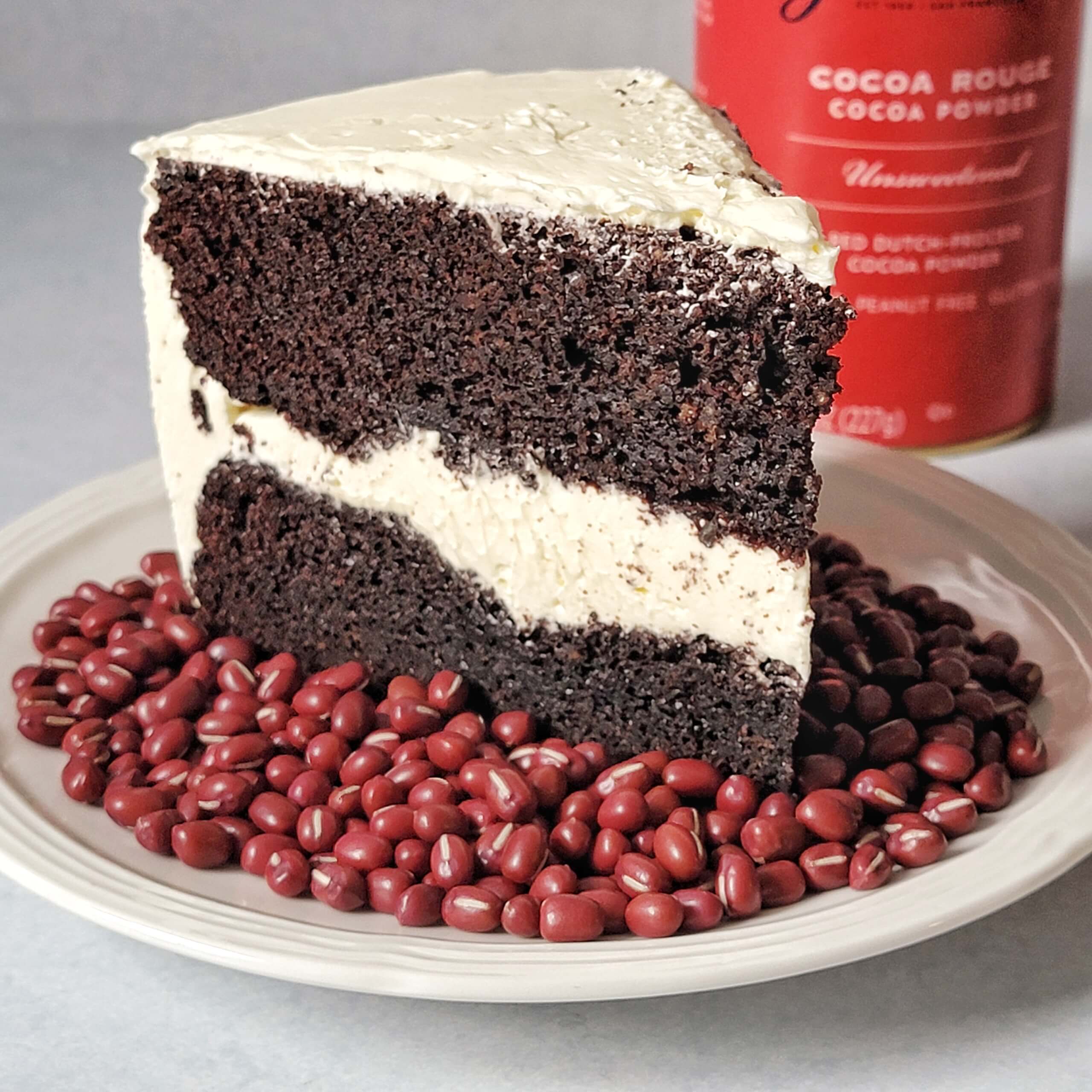 Photo of a thick slice of rich chocolate layer cake with white frosting on a plate surrounded by dry adzuki beans. A container of cocoa powder is in the background.