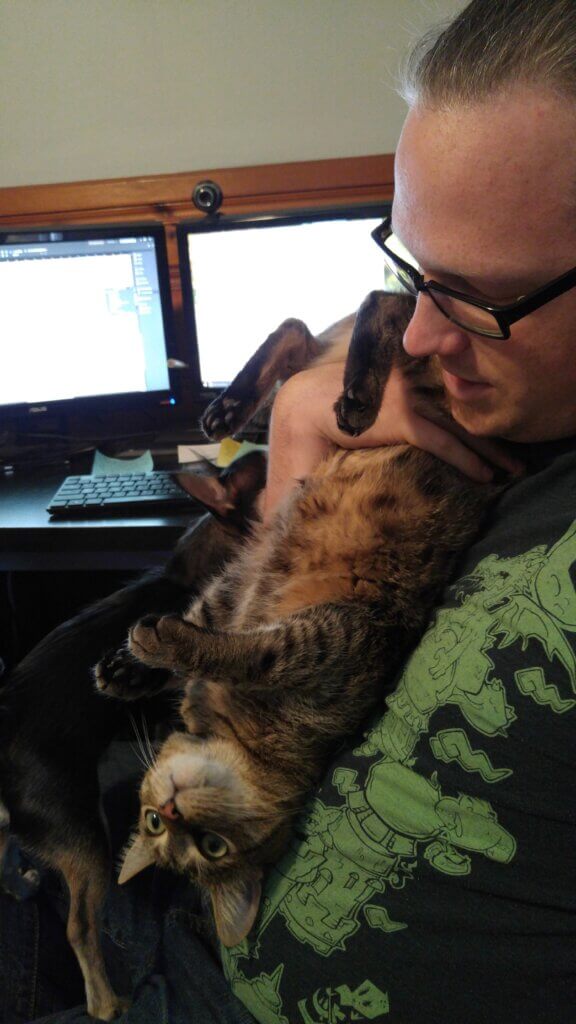Cory sits at the computer holding Loki to his chest upside down. Loki appears to be fine with this.