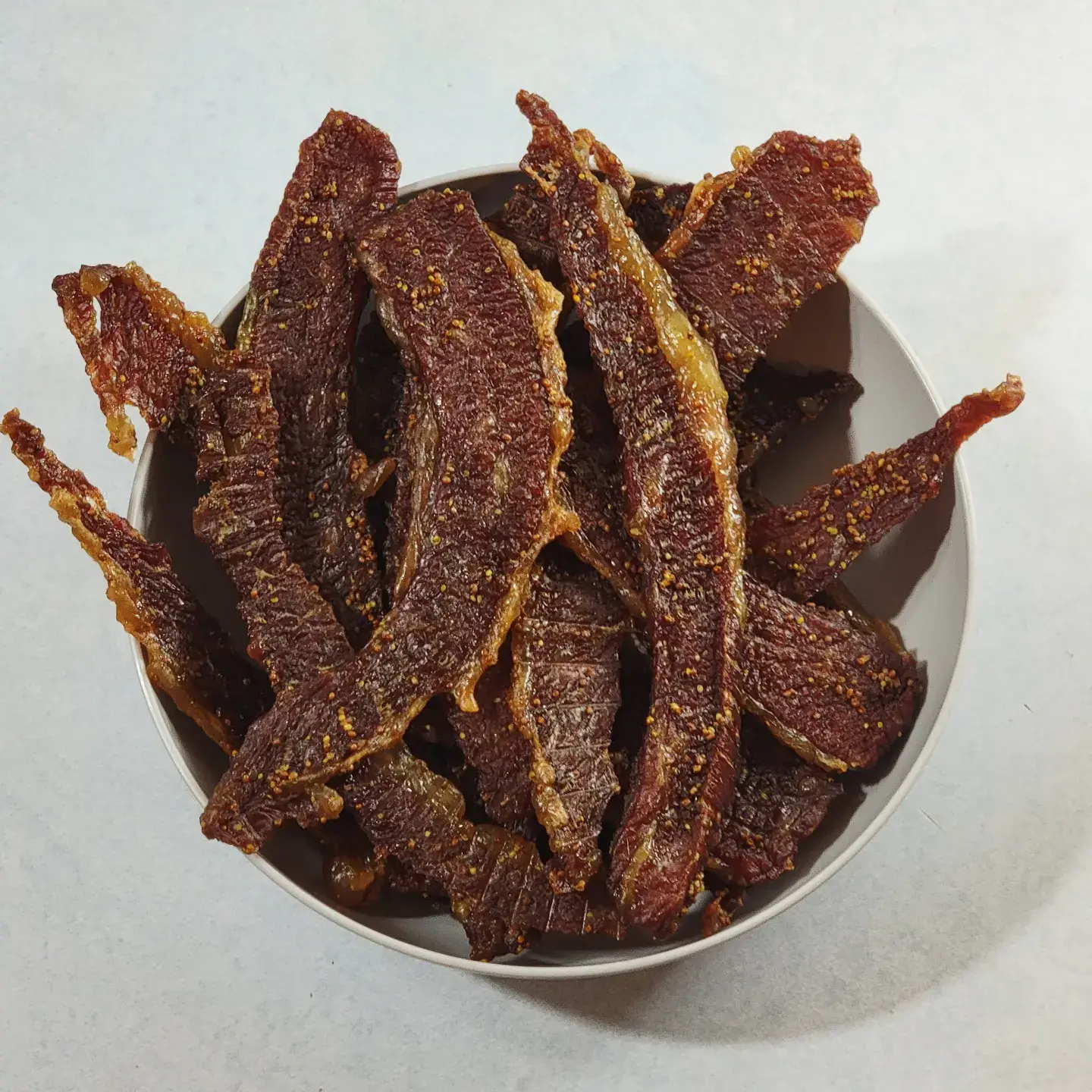 Bowl filled to the brim with corned beef jerky. The color is a deep maroon with a slightly shiny surface dotted with mustard seeds. On most of the slices, there is a gleaming rind of of fat.