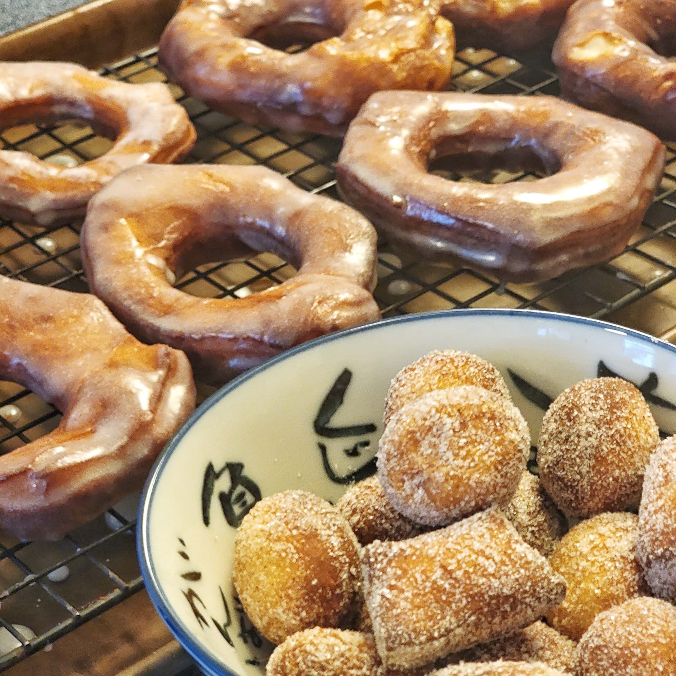 Freshly-glazed donuts resting on a wire rack. A bowl of sugar-coated donut holes is in the foreground.