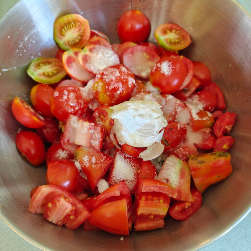 Chopped paste tomatoes in a large metal bowl topped with smashed garlic, salt, and a thin white paste (shio koji).